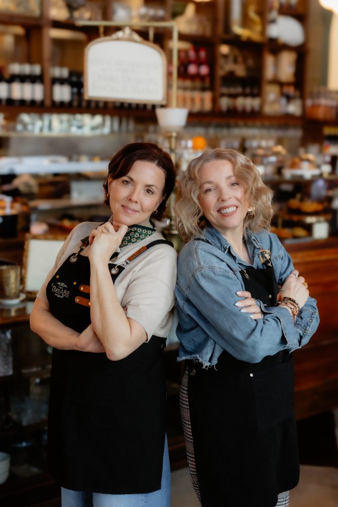 Two cafe owners smiling back-to-back indoors